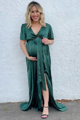 woman wearing a dark green satin maternity gown with a tie waist and a side slit, holding her baby bump