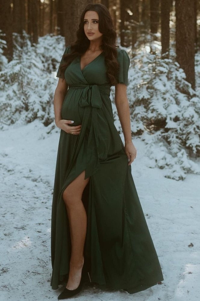 woman wearing a green maternity maxi dress with a wrap design, showing a baby bump in a snowy forest setting