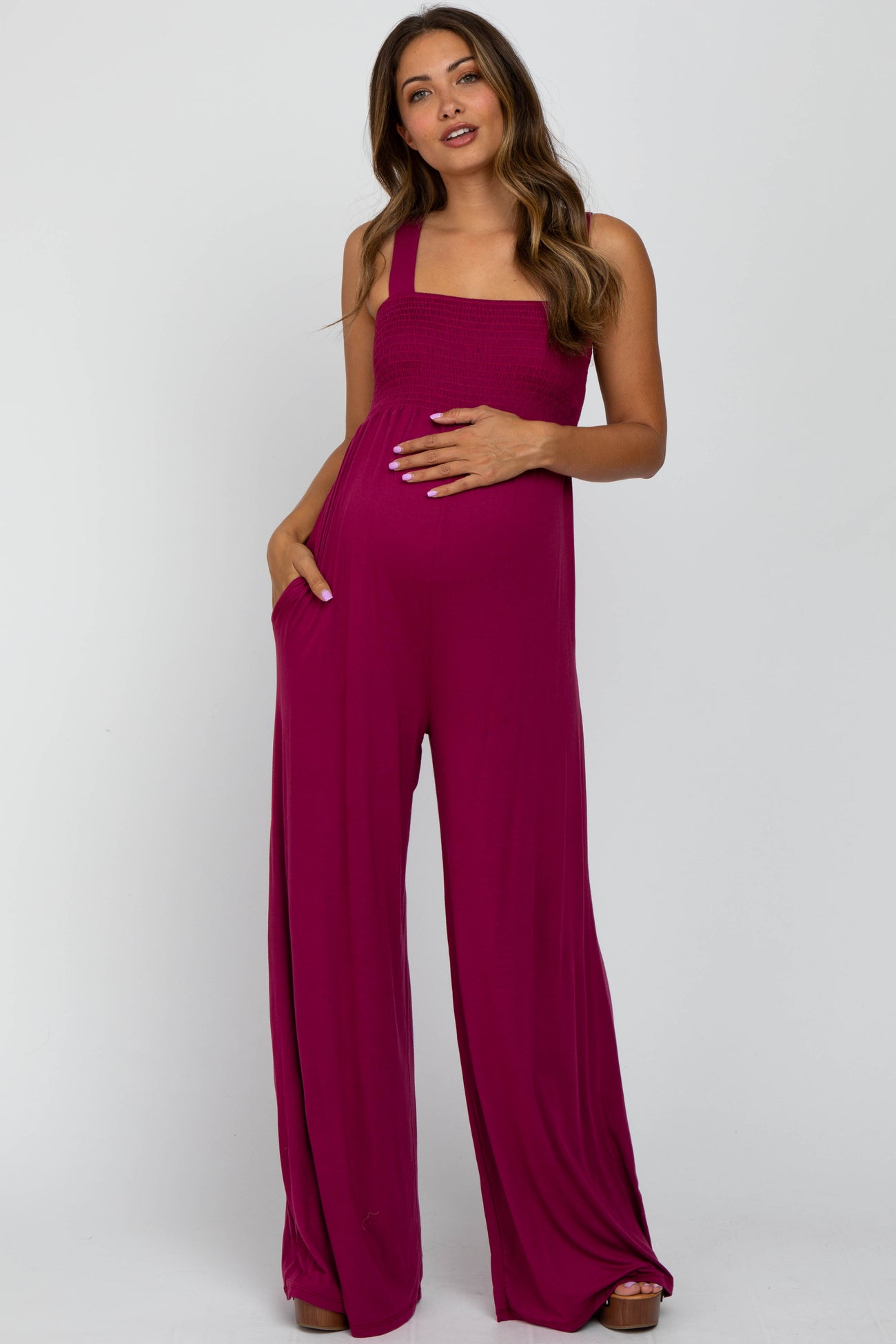 woman wearing a fuchsia maternity jumpsuit with a smocked bodice and side pockets, holding her baby bump