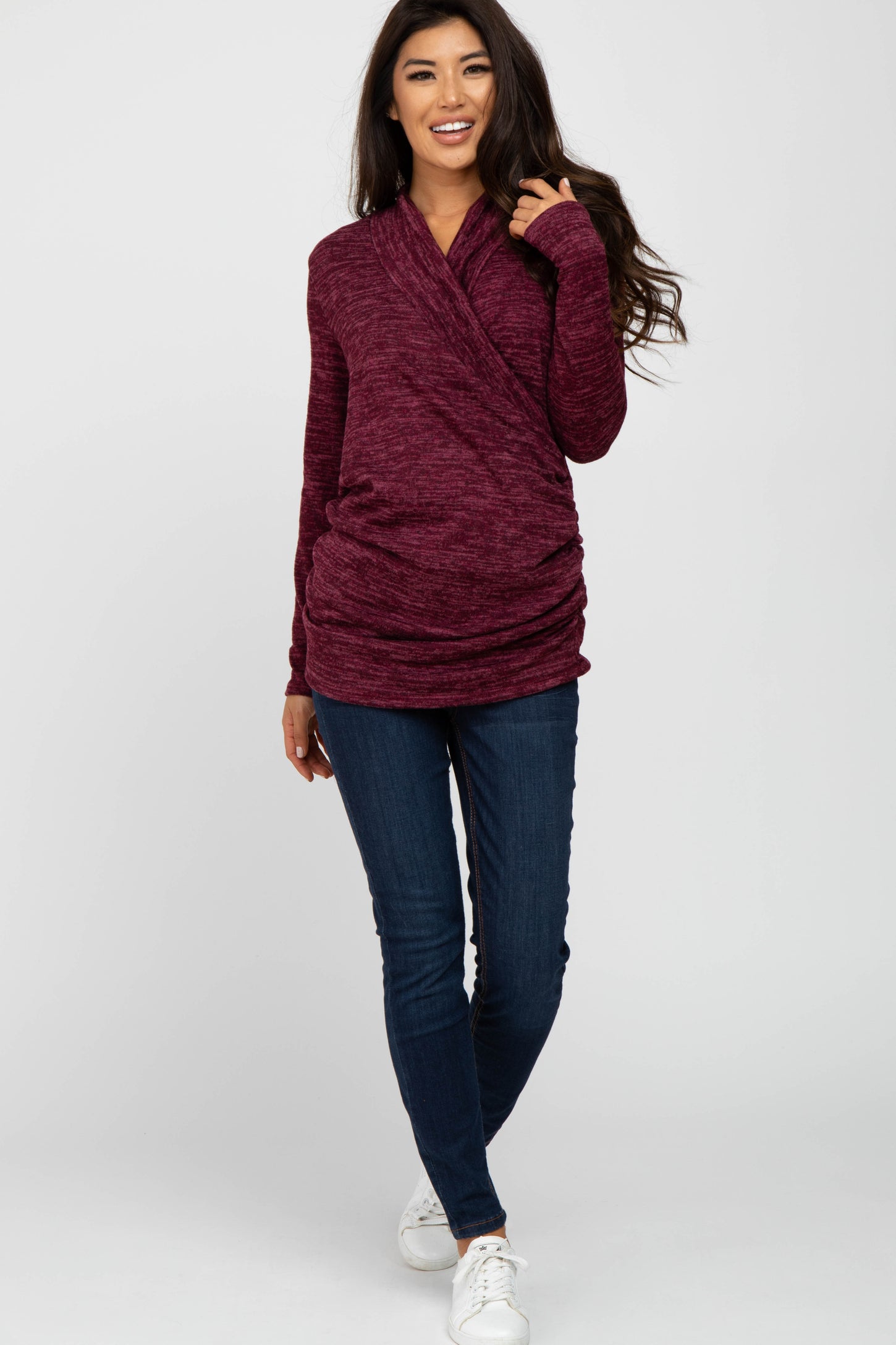 woman wearing a burgundy long sleeve wrap top with draped details and blue skinny jeans, smiling at the camera