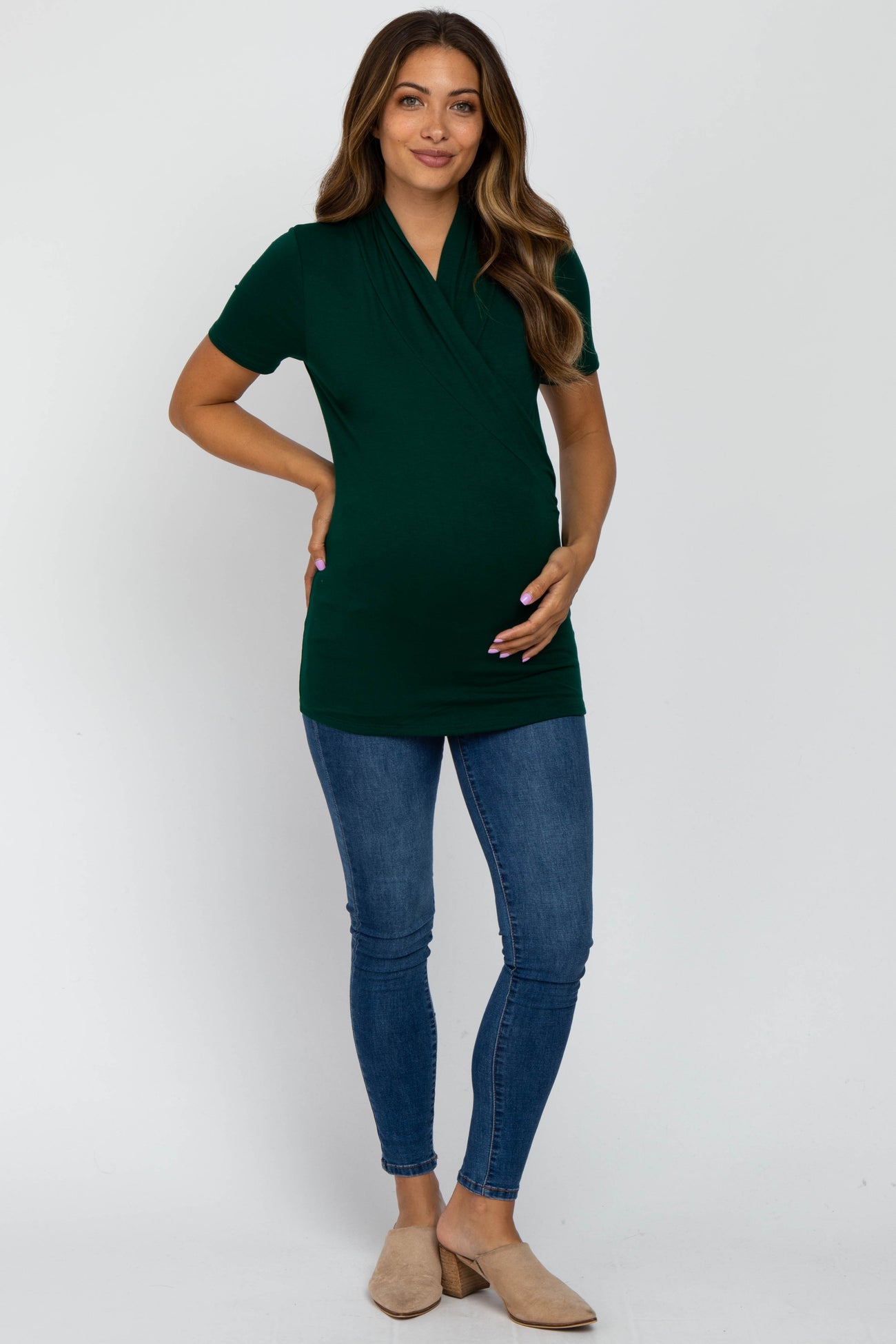 woman wearing a dark green short sleeve maternity top and holding her baby bump with blue skinny jeans