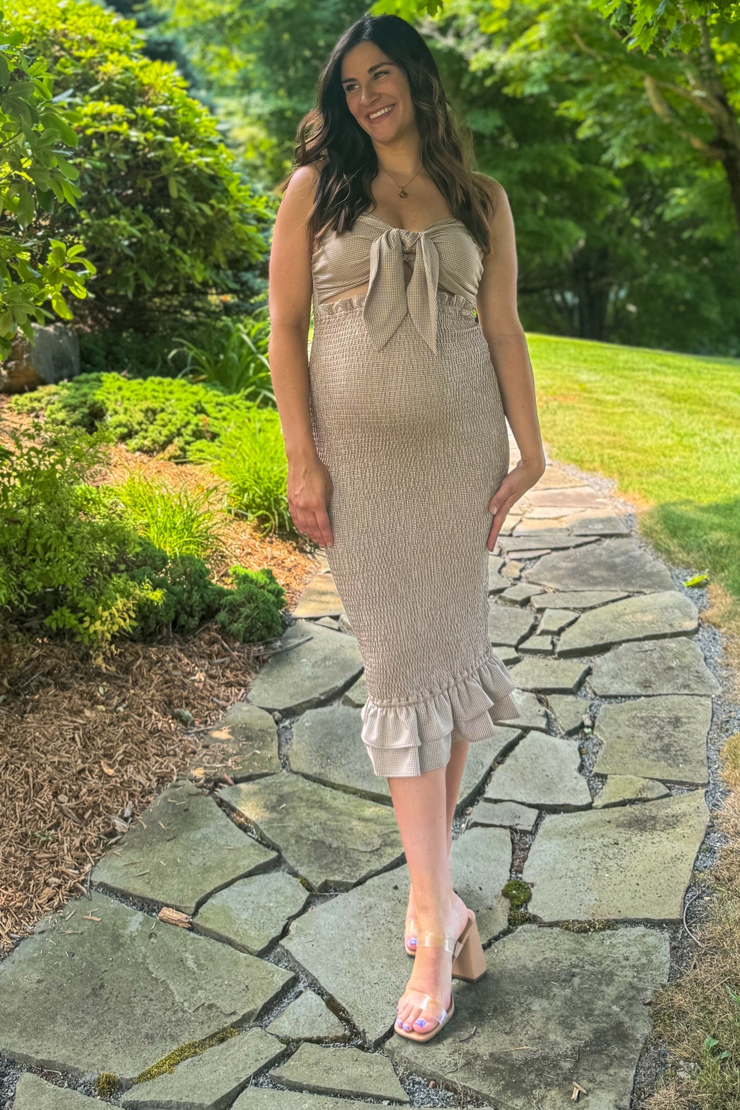 woman wearing a beige maternity dress with a bow detail and ruffle hem, showing her baby bump while walking on a stone path
