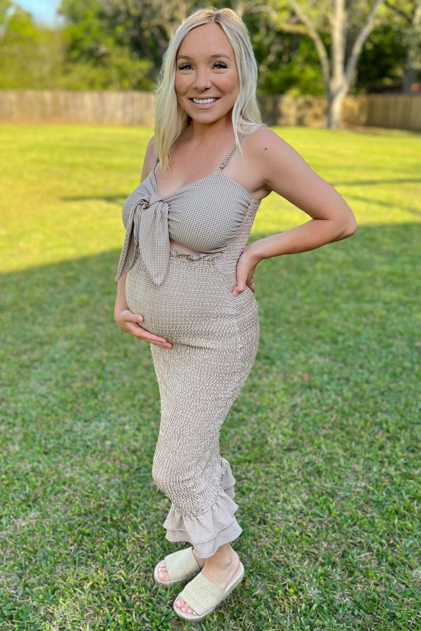 woman wearing a light beige maternity dress with a tie front and ruffled hem, showing her baby bump while standing outdoors