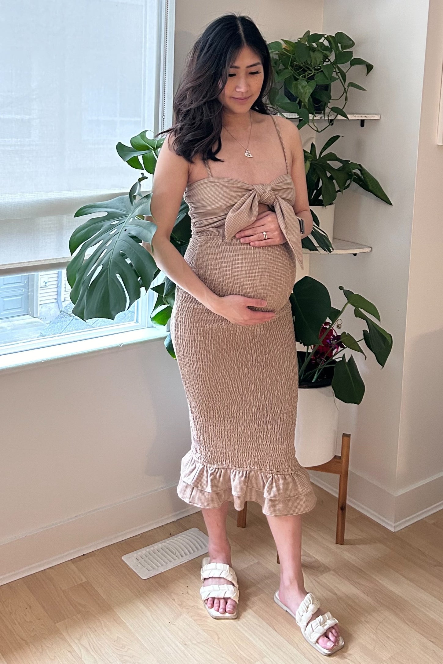 woman wearing a beige maternity dress with a bow detail and ruffle hem, holding her baby bump