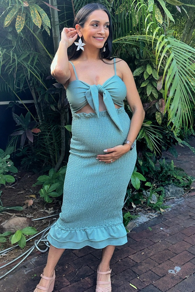 woman wearing a teal maternity dress with a tie front and ruffle hem, holding her baby bump amidst lush greenery
