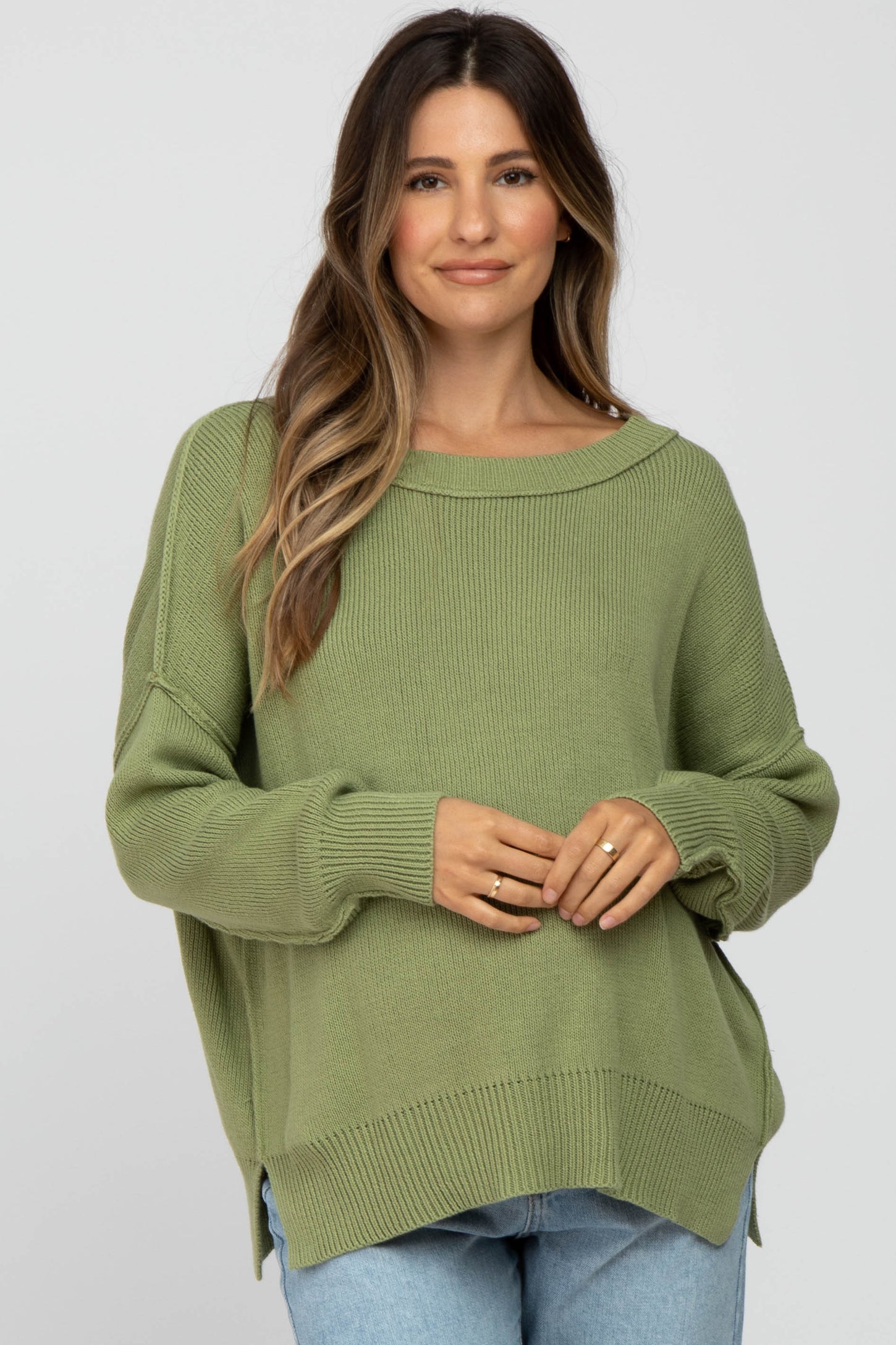 woman wearing a light green oversized maternity sweater with a boat neckline and side slits, holding her baby bump