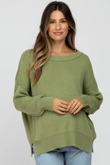 woman wearing a light green oversized maternity sweater with a boat neckline and side slits, holding her baby bump