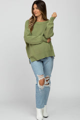 woman wearing a light green maternity sweater with a relaxed fit and distressed jeans, smiling and posing with one hand on her hip