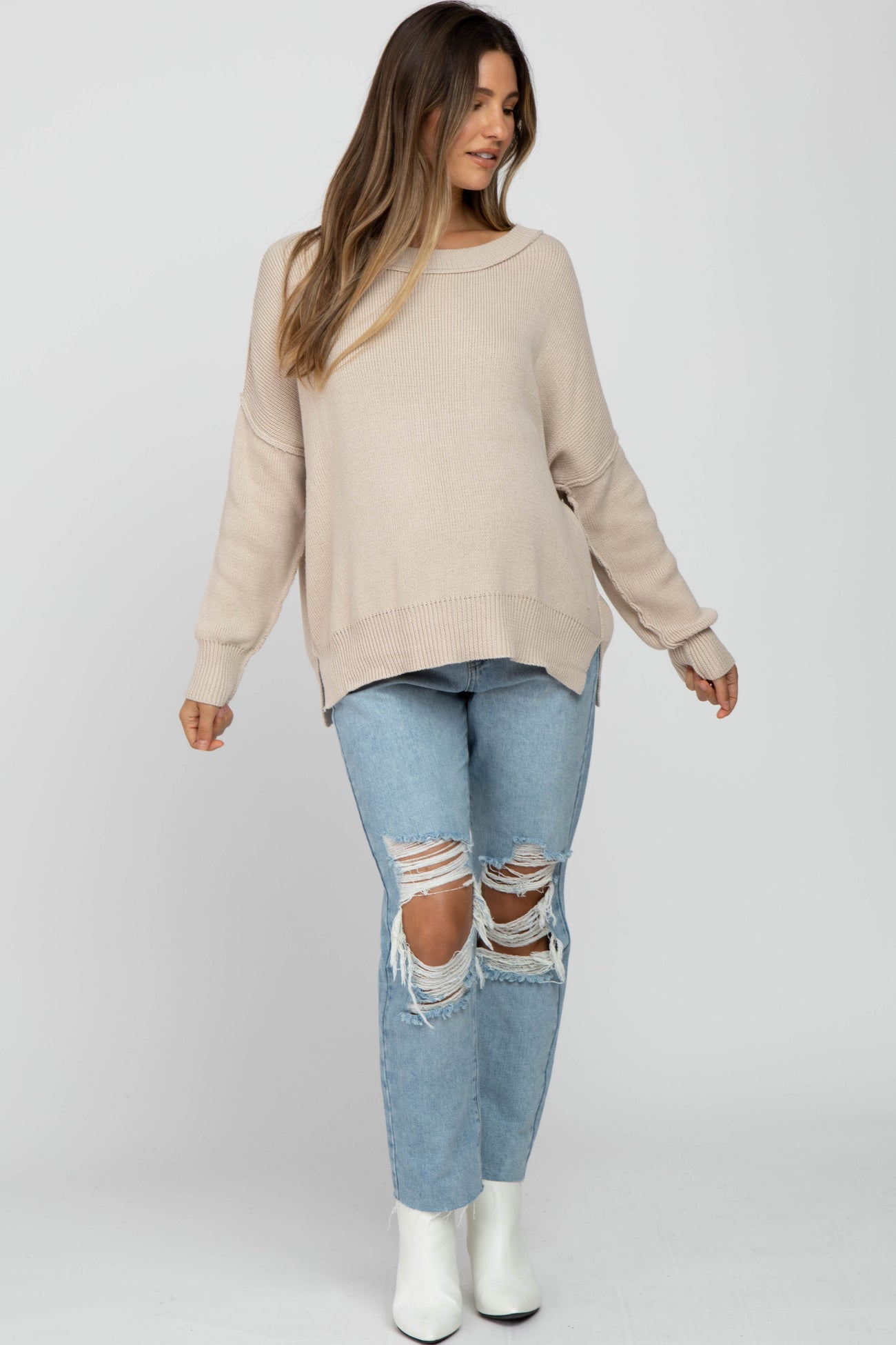 woman wearing a beige oversized maternity sweater and ripped light blue jeans, showing a baby bump