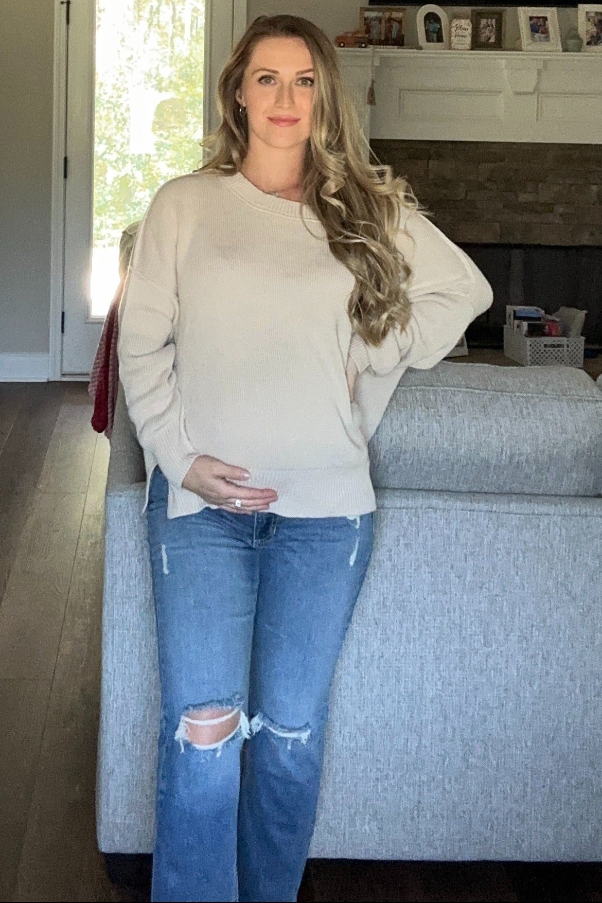 woman wearing a light beige maternity sweater and distressed blue jeans, holding her baby bump while standing by a couch