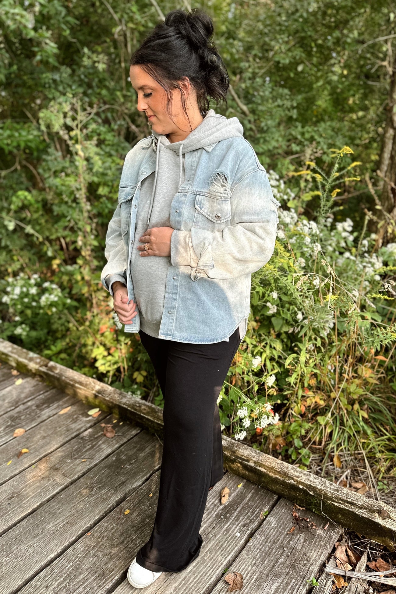 woman wearing a light blue denim jacket over a gray hoodie and black maternity pants, showing her baby bump
