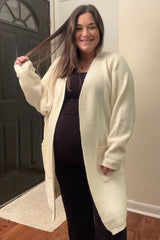 woman wearing a cream long sleeve maternity cardigan over a black dress and displaying her baby bump