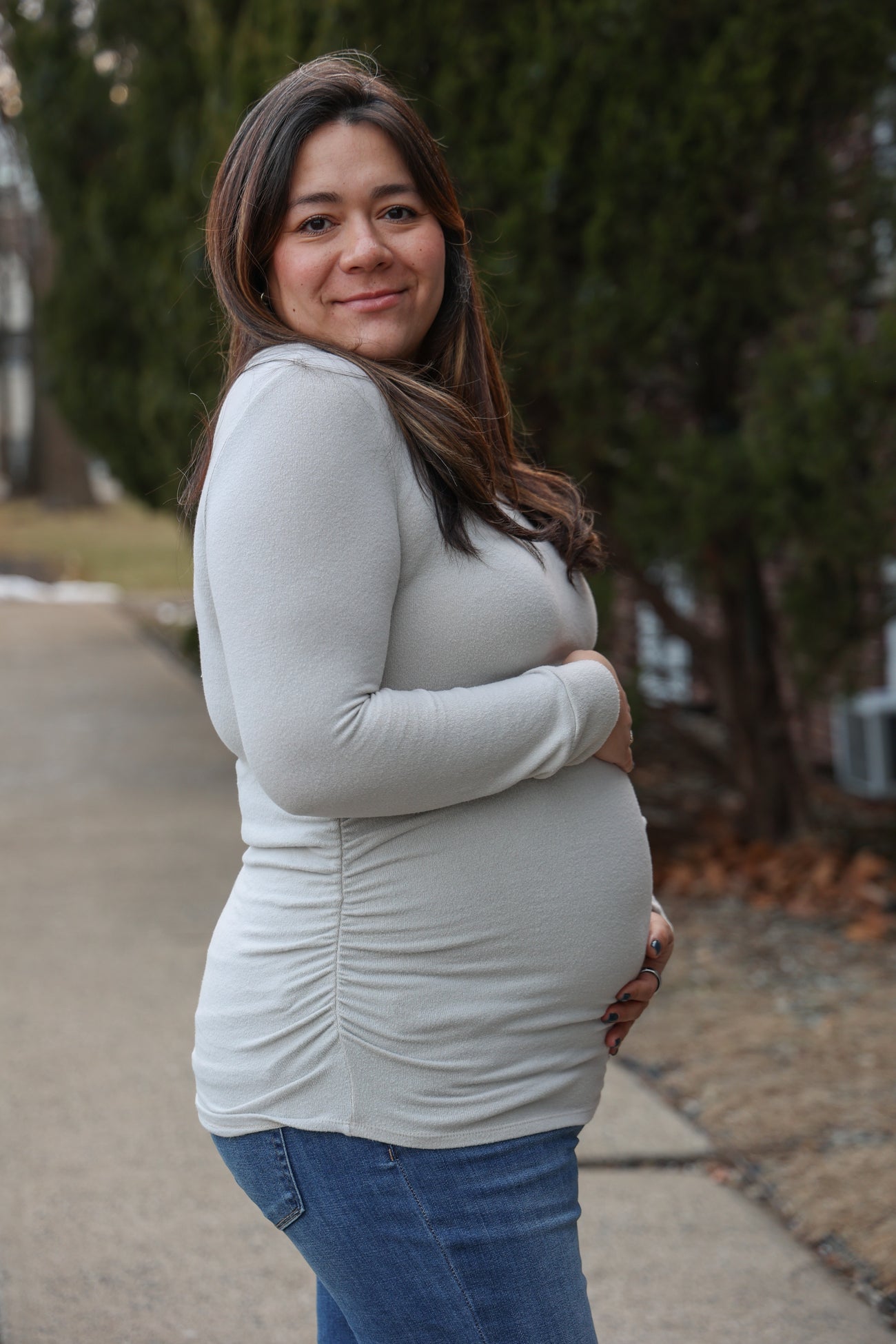 woman wearing a light grey long sleeve maternity top and holding her baby bump while standing outdoors