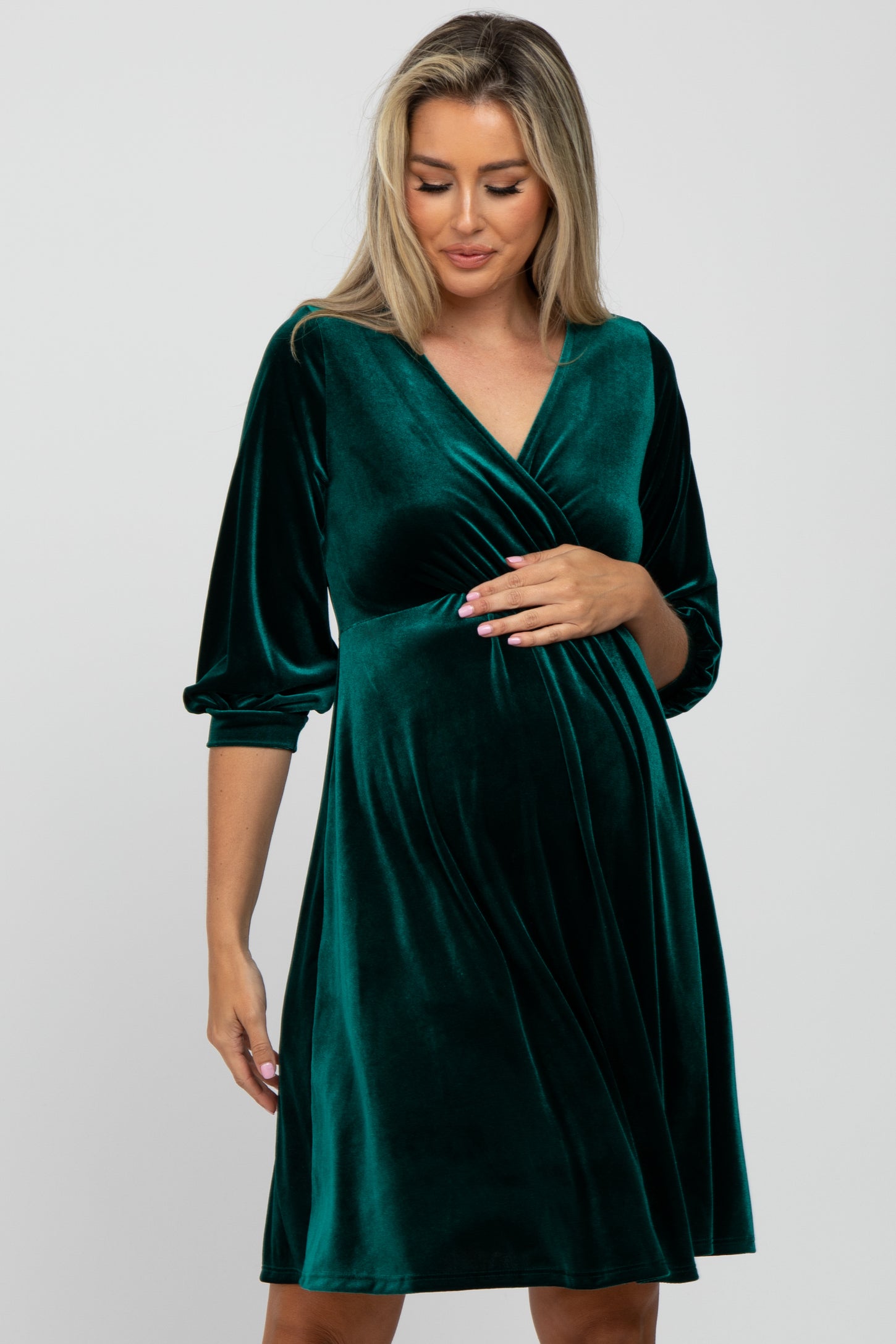 woman wearing a deep green velvet maternity dress with three-quarter sleeves and a wrap style, cradling her baby bump