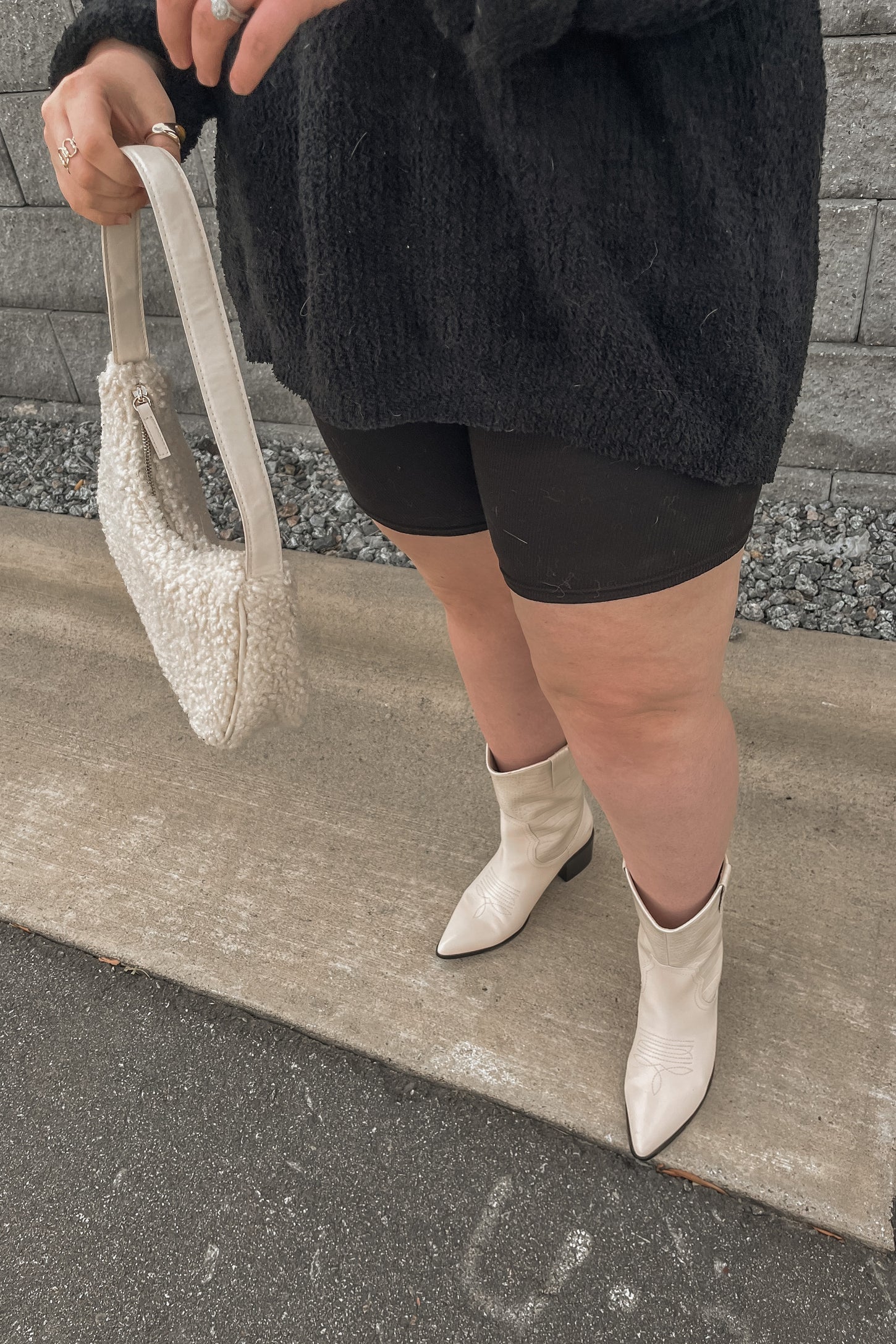 woman wearing a black oversized sweater, black shorts, and white cowboy boots, holding a cream-colored bag