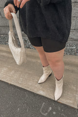 woman wearing a black oversized sweater, black shorts, and white cowboy boots, holding a cream-colored bag