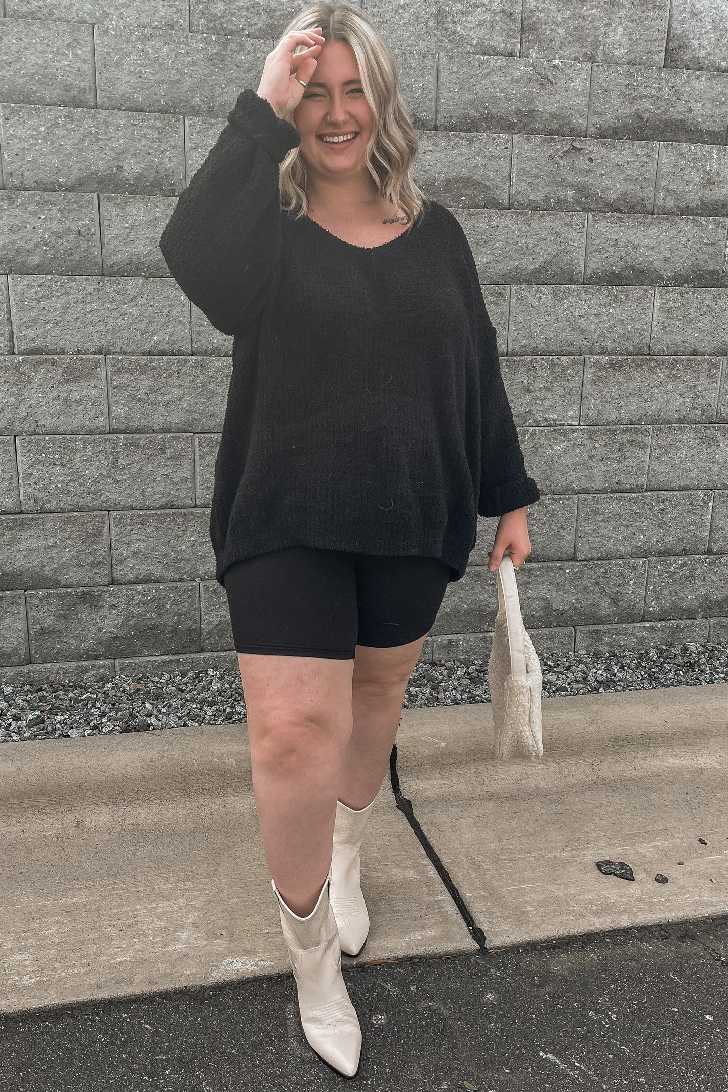 woman wearing an oversized black sweater with black bike shorts and white knee-high boots, holding a small white bag