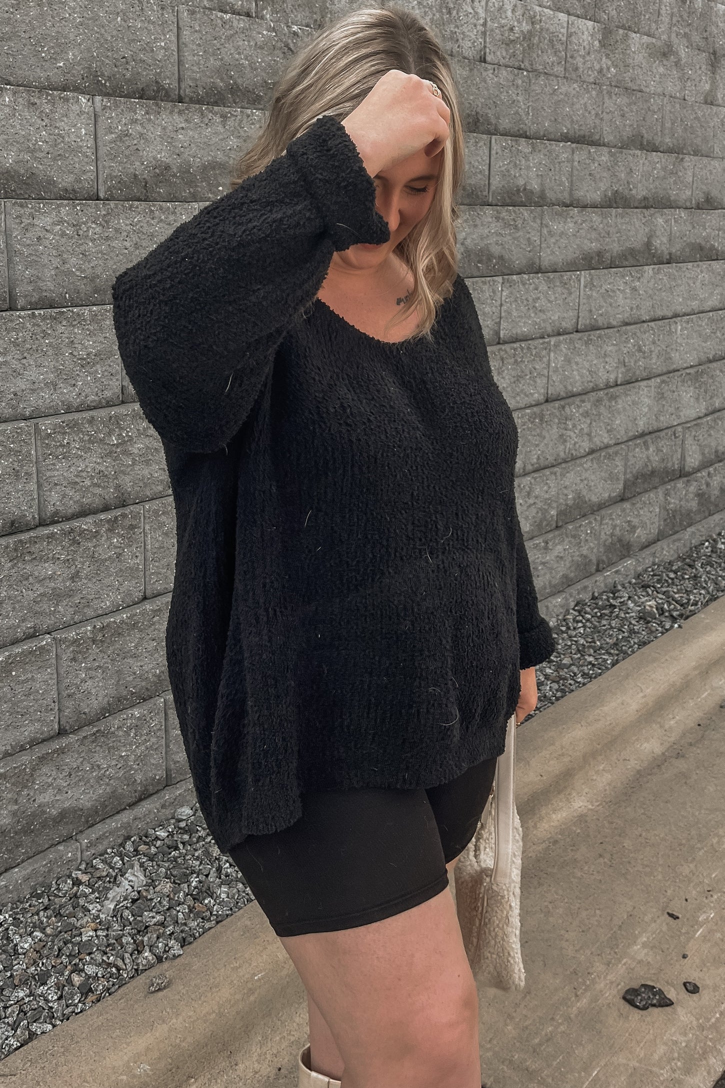 woman wearing a black oversized textured sweater and black shorts, standing outdoors against a gray wall