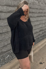 woman wearing a black oversized textured sweater and black shorts, standing outdoors against a gray wall