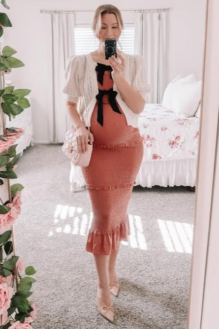 pregnant woman in pink smocked maternity dress and white cardigan with black bows taking mirror selfie