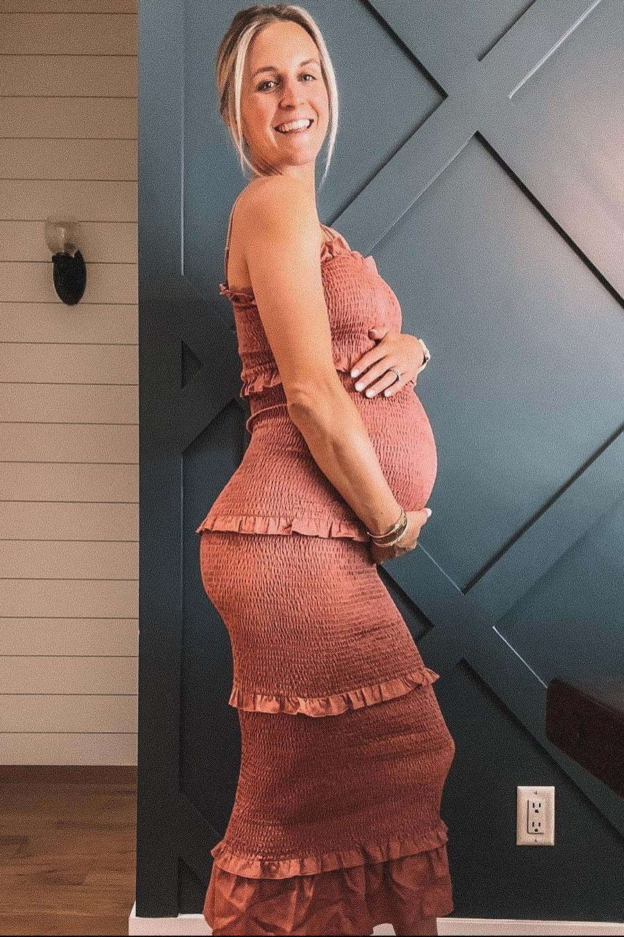 woman wearing a rose-colored maternity dress with ruffles, showing her baby bump and standing against a patterned wall