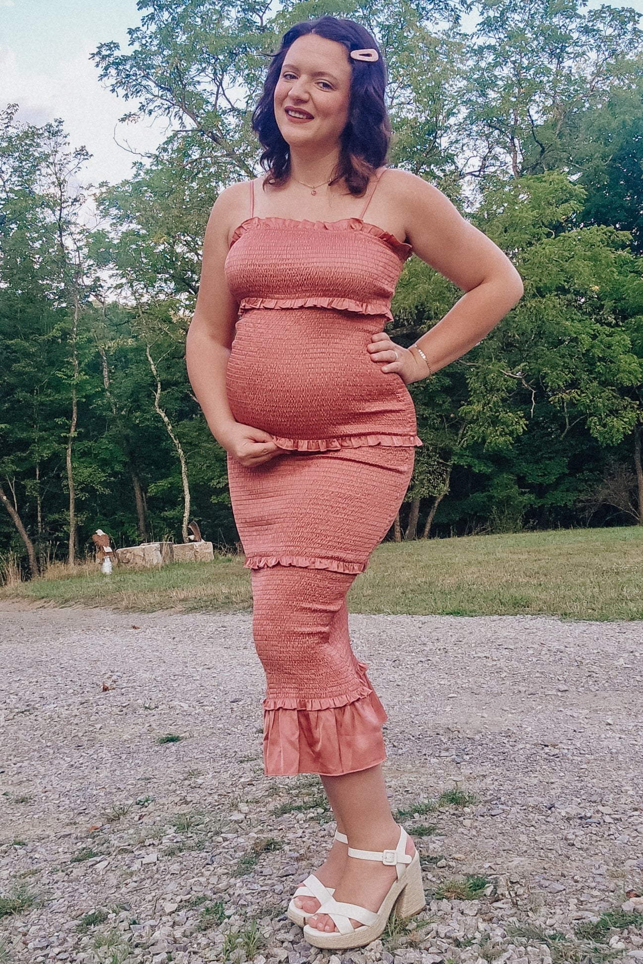 woman wearing a pink ruffled maternity dress and holding her baby bump in an outdoor setting