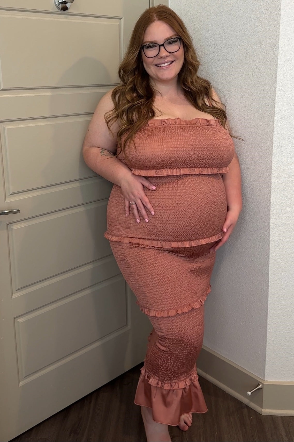 woman wearing a maternity peach smocked dress with ruffles and a visible baby bump, smiling against a door