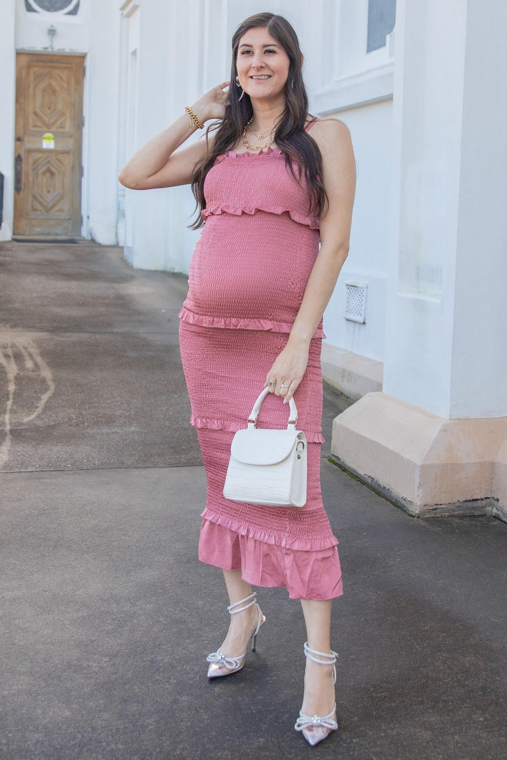 Pregnant woman in a pink smocked maternity dress holding a white handbag and wearing silver heels