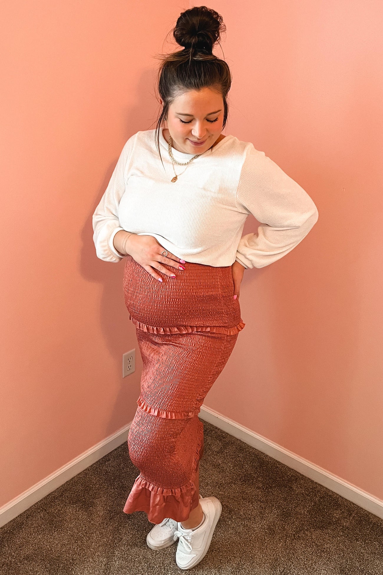 woman wearing a white long sleeve top and a rust-colored maternity skirt, showing her baby bump while posing against a pink wall