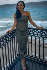 woman wearing a khaki green ruffled maternity dress, standing on a balcony with a beach backdrop and showing her baby bump