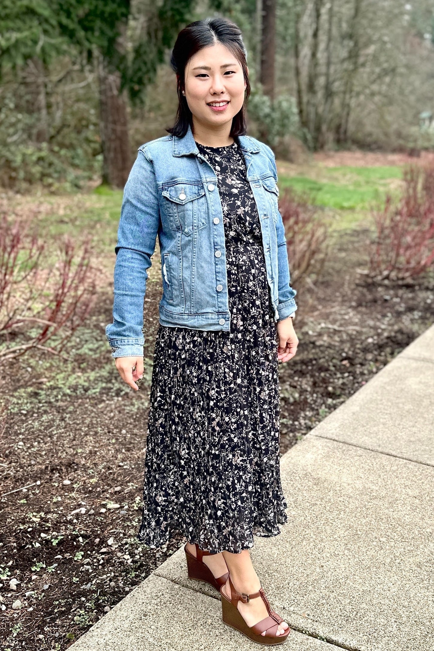 woman wearing a black floral maxi dress, denim jacket, and brown wedge sandals, standing outdoors