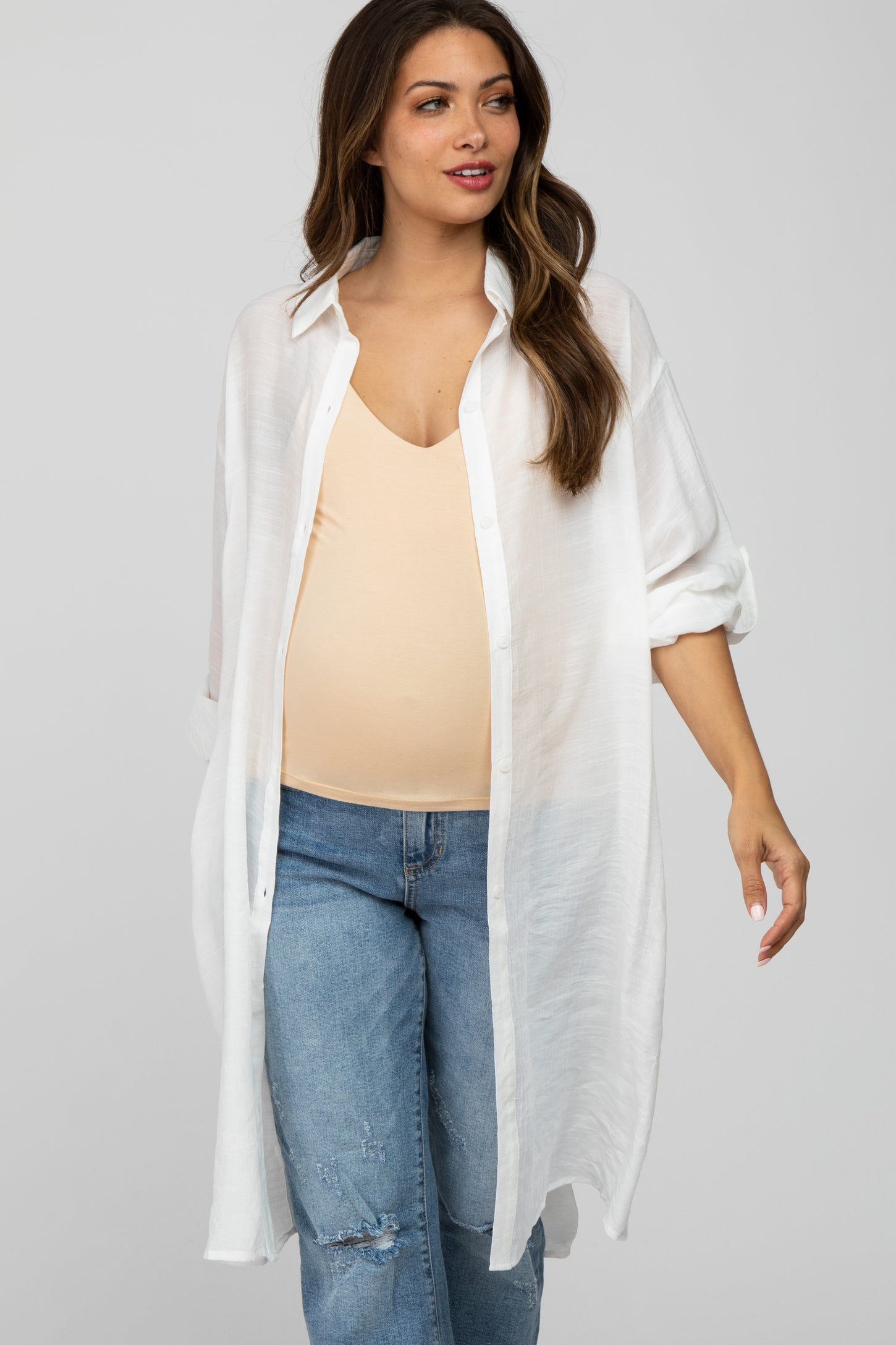 woman wearing a white oversized button-up maternity shirt over a light beige tank top and distressed jeans, showing her baby bump