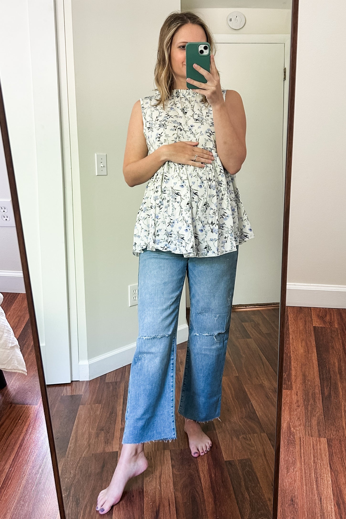 woman wearing a floral sleeveless maternity top and distressed blue jeans, holding her baby bump in front of a mirror