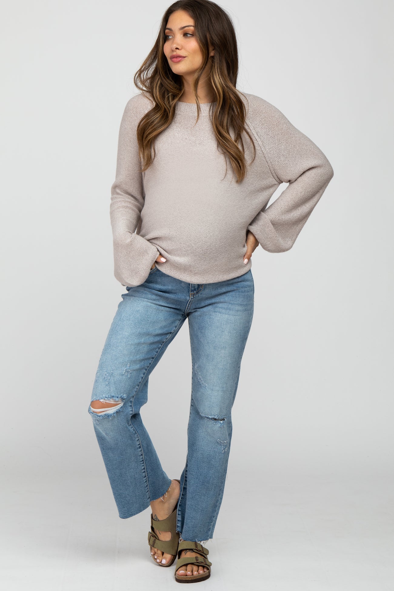 woman wearing a light beige oversized maternity sweater and distressed blue jeans, showing her baby bump