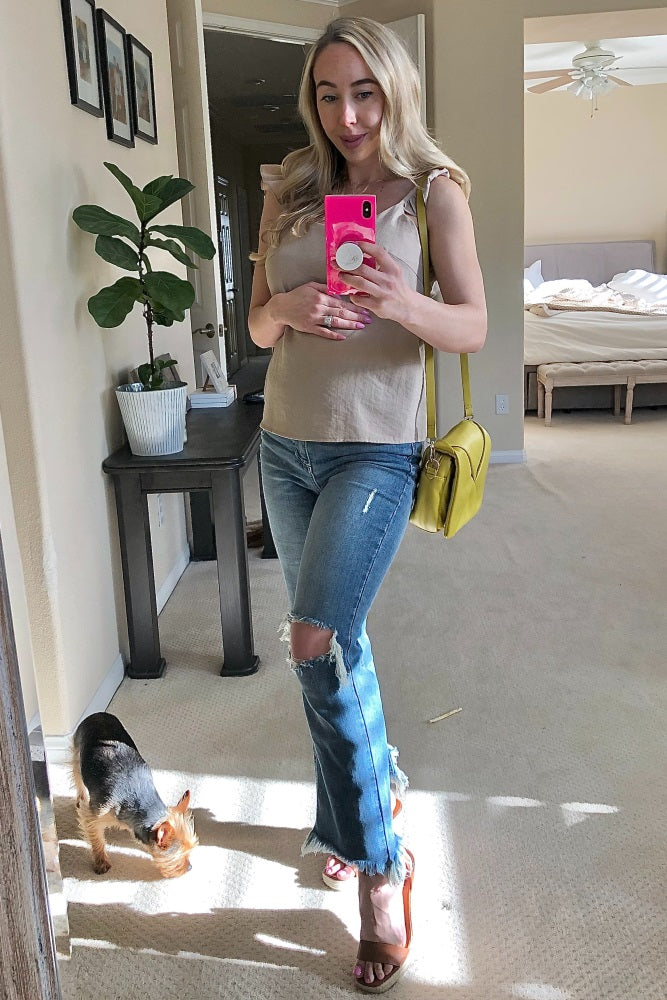 Woman in tan top and distressed jeans taking mirror selfie with pink phone and yellow crossbody bag