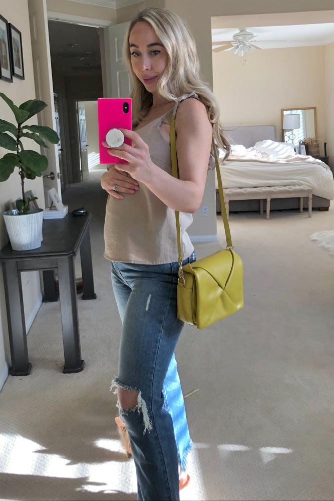woman wearing a beige maternity tank top and distressed blue jeans, holding a yellow handbag and showing her baby bump