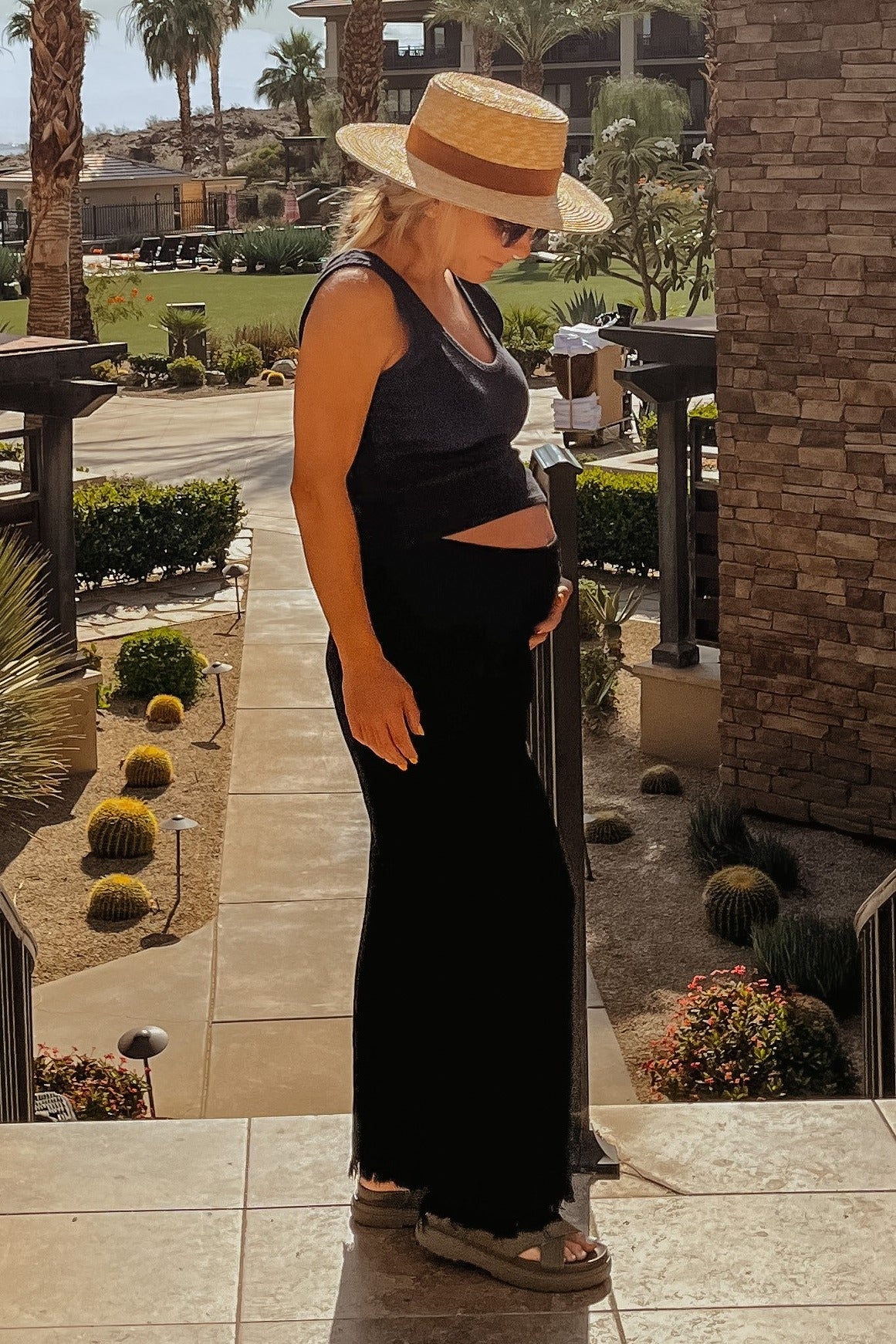 woman wearing a black maternity maxi dress and a sun hat, gently holding her baby bump while standing outdoors