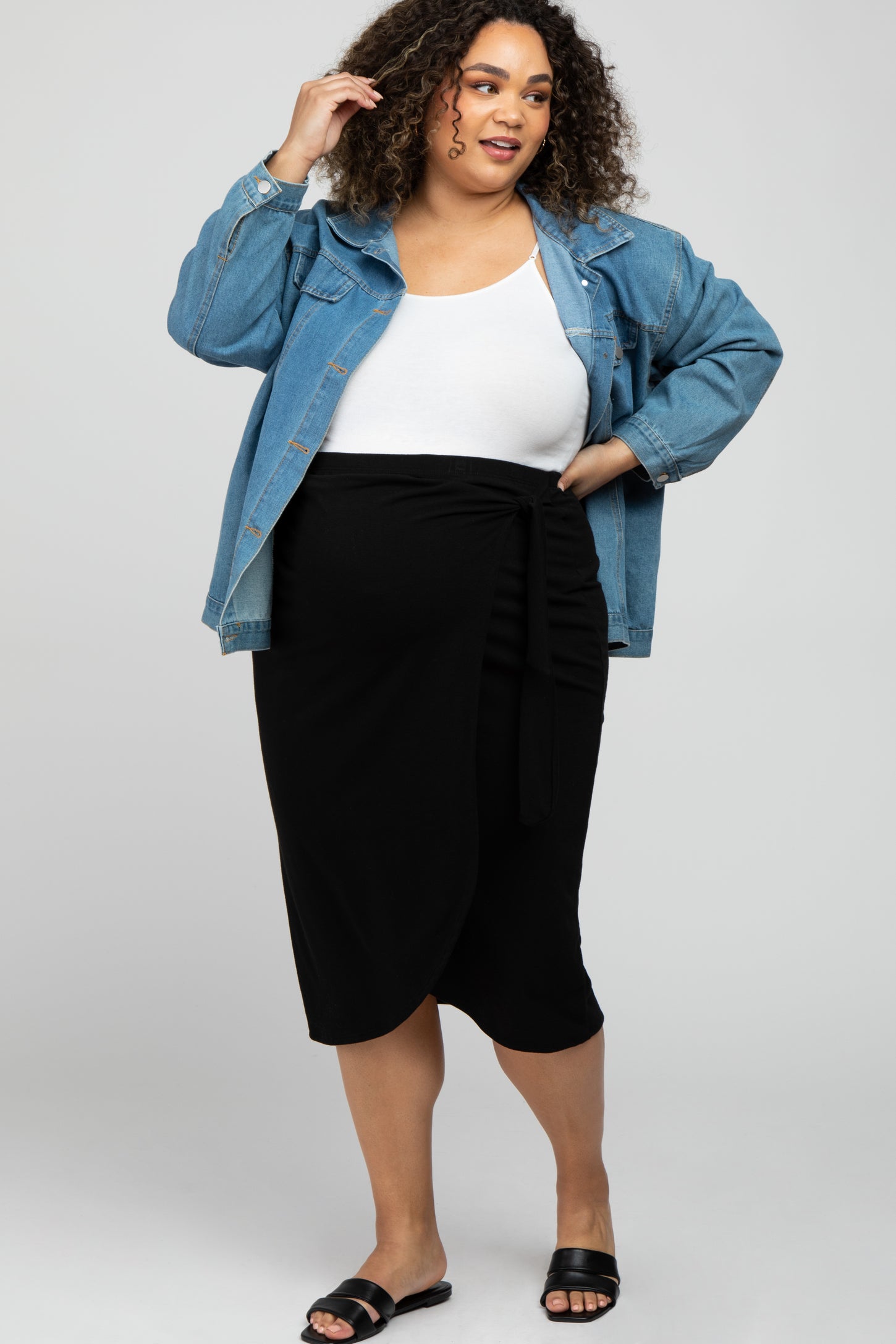 Plus size woman in denim jacket, white tank top, and black wrap midi skirt with sandals