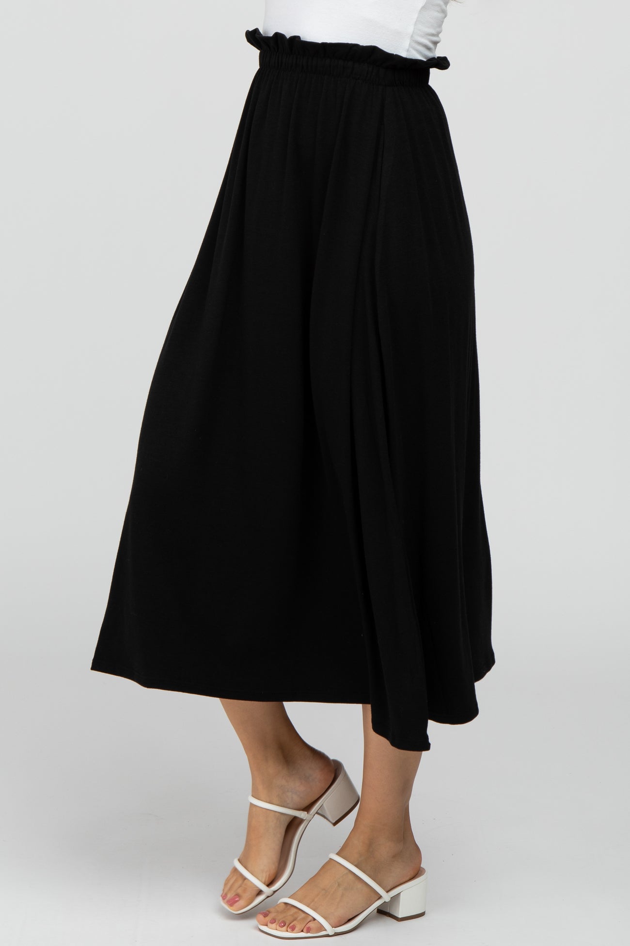 Woman wearing a black flowy midi skirt with a ruffled elastic waistband and white strappy heeled sandals
