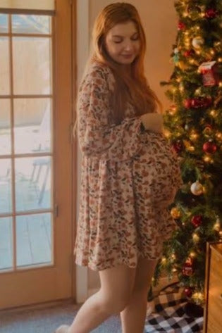 woman wearing a floral maternity dress, gently cradling her baby bump in front of a decorated Christmas tree