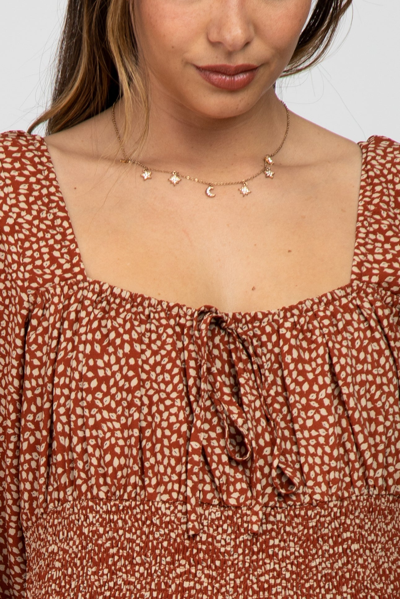 woman wearing a rust-colored maternity blouse with a square neckline and subtle floral print, featuring a tie detail and short puff sleeves