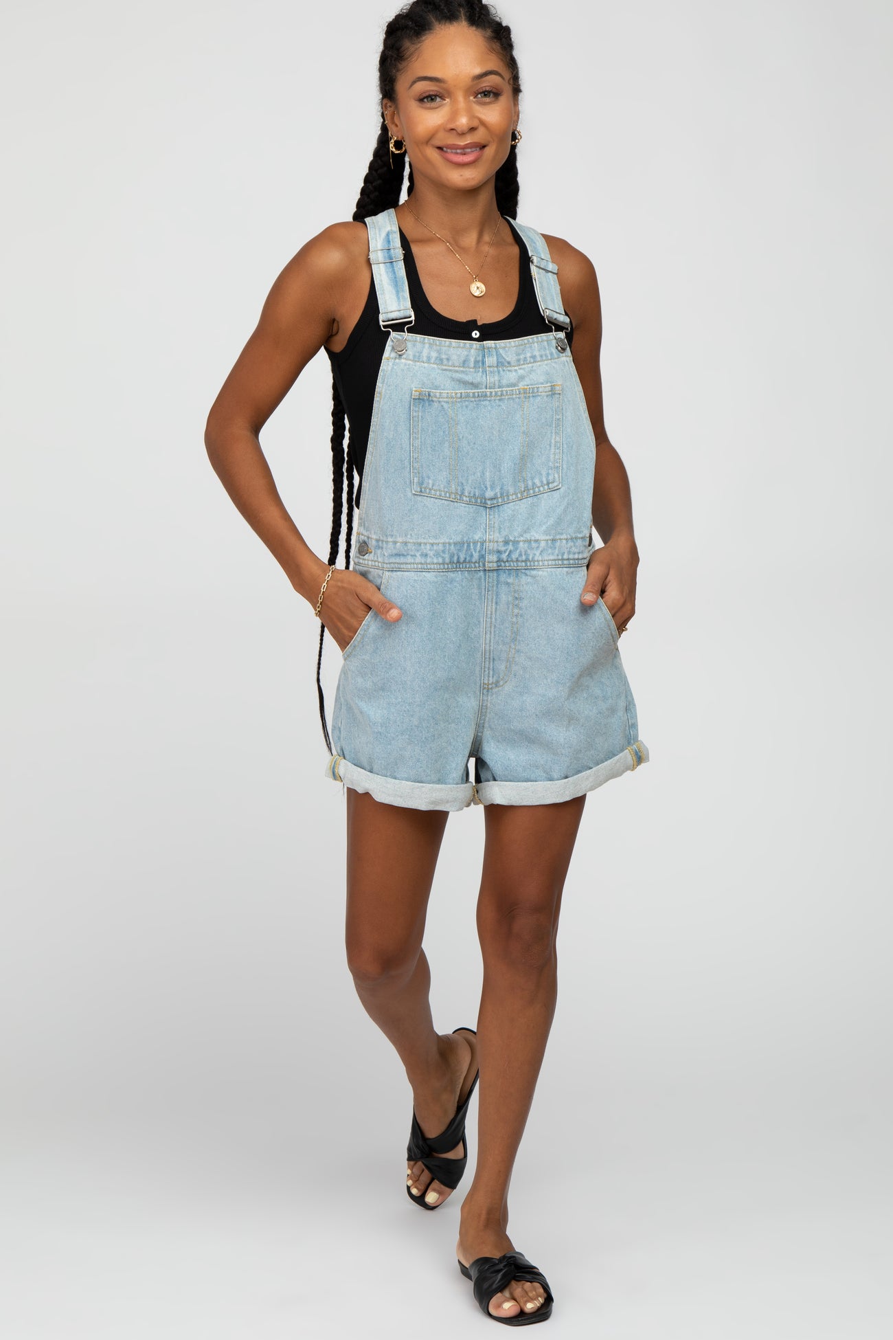woman wearing a light wash denim overall shorts with rolled cuffs and a black tank top, standing and smiling