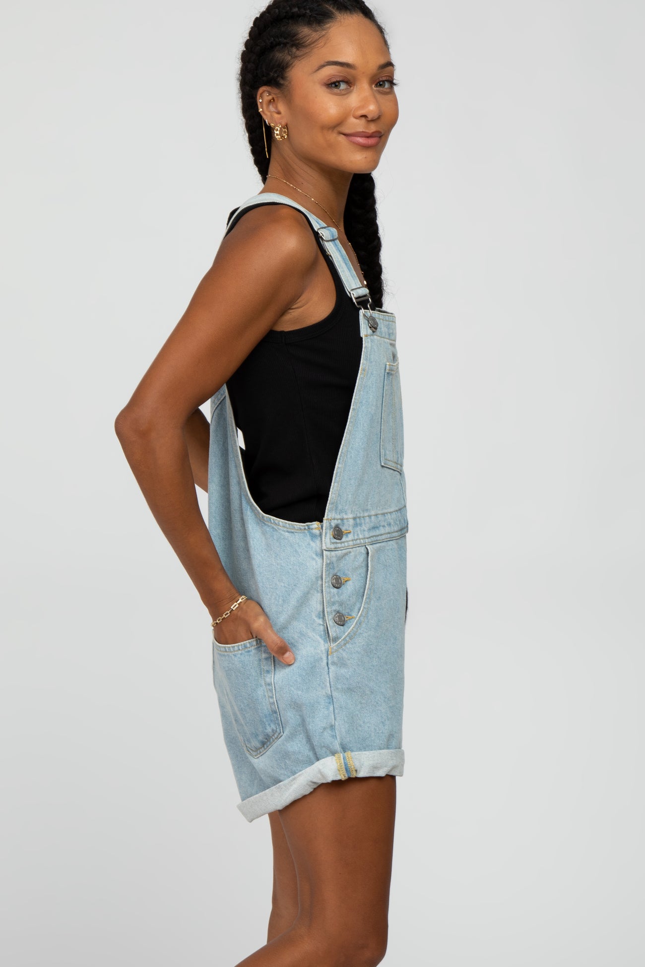 woman wearing light wash denim short overalls and a black tank top against a white background