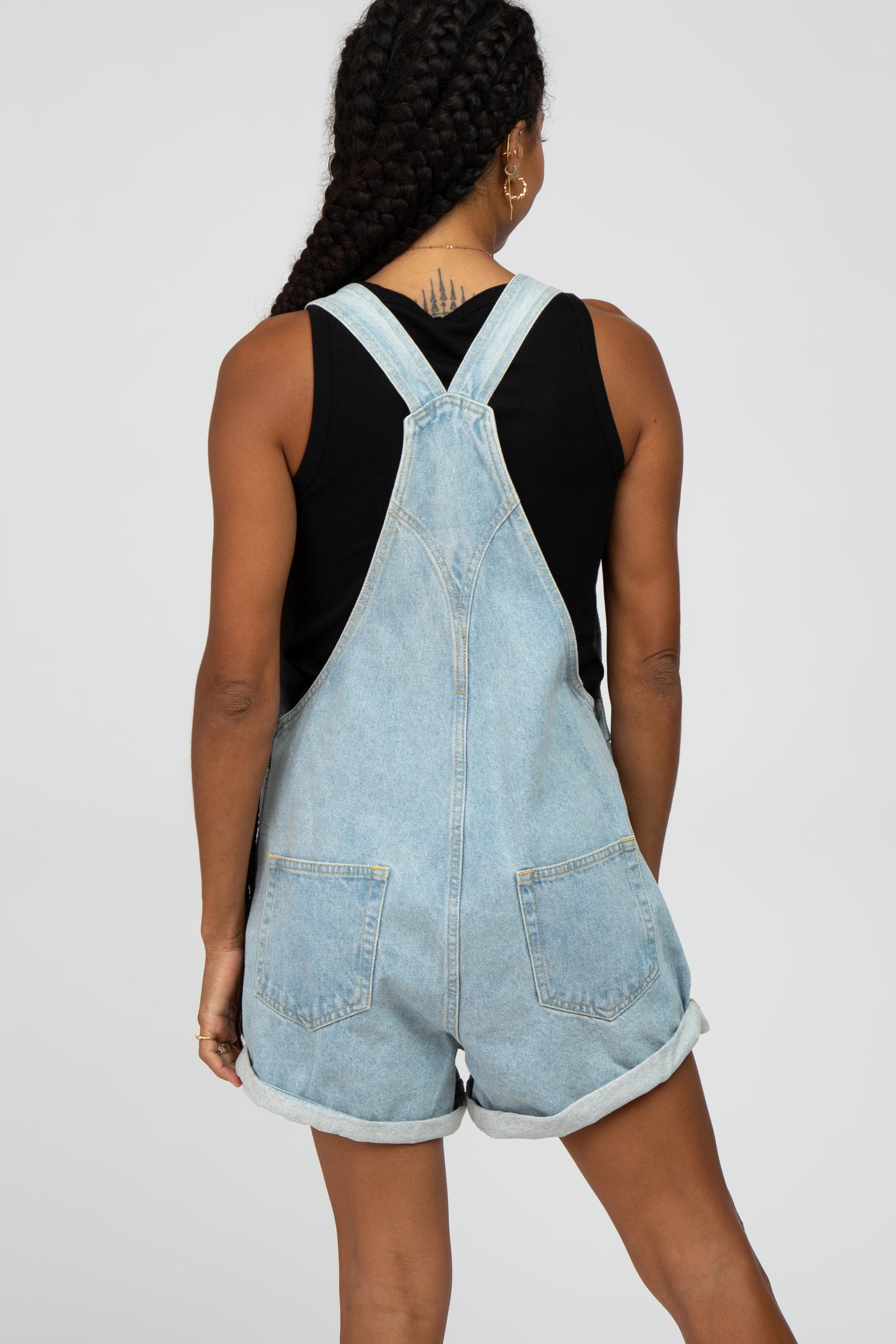 woman wearing a light wash denim overall shorts with rolled hems and a black sleeveless top, seen from the back