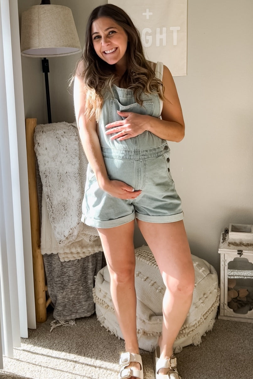 woman wearing a light green maternity overalls and holding her baby bump, standing indoors with a cozy background