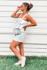woman wearing a light blue maternity overall shorts and a white tank top, showing her baby bump and styled with sunglasses