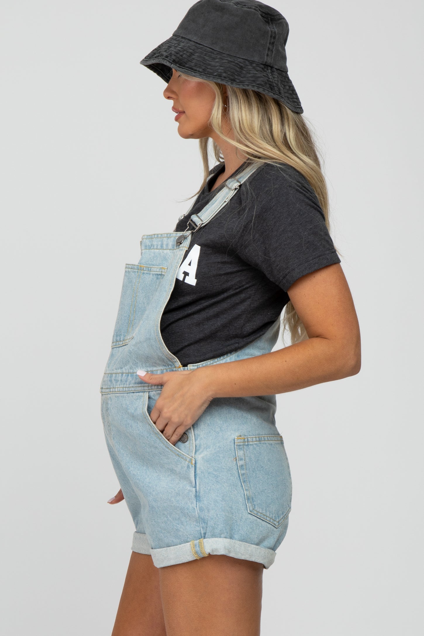 woman wearing light blue denim overalls with rolled hem and pockets, paired with a gray t-shirt and black bucket hat, showing baby bump