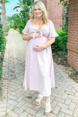 woman wearing a pink maternity dress with tie detail holding her baby bump outdoors
