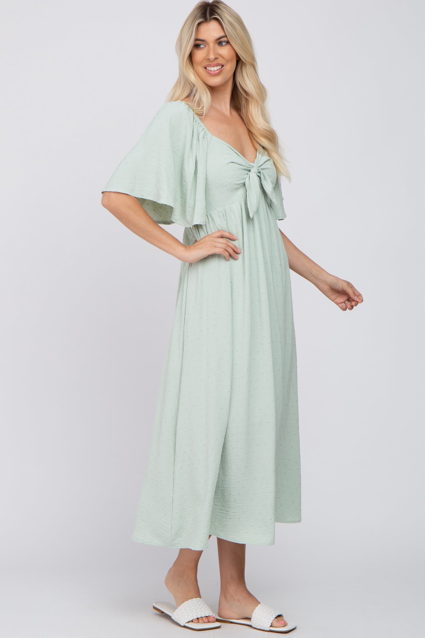 woman wearing a mint green flutter sleeve maxi dress with a front knot detail and styled with white slip-on sandals
