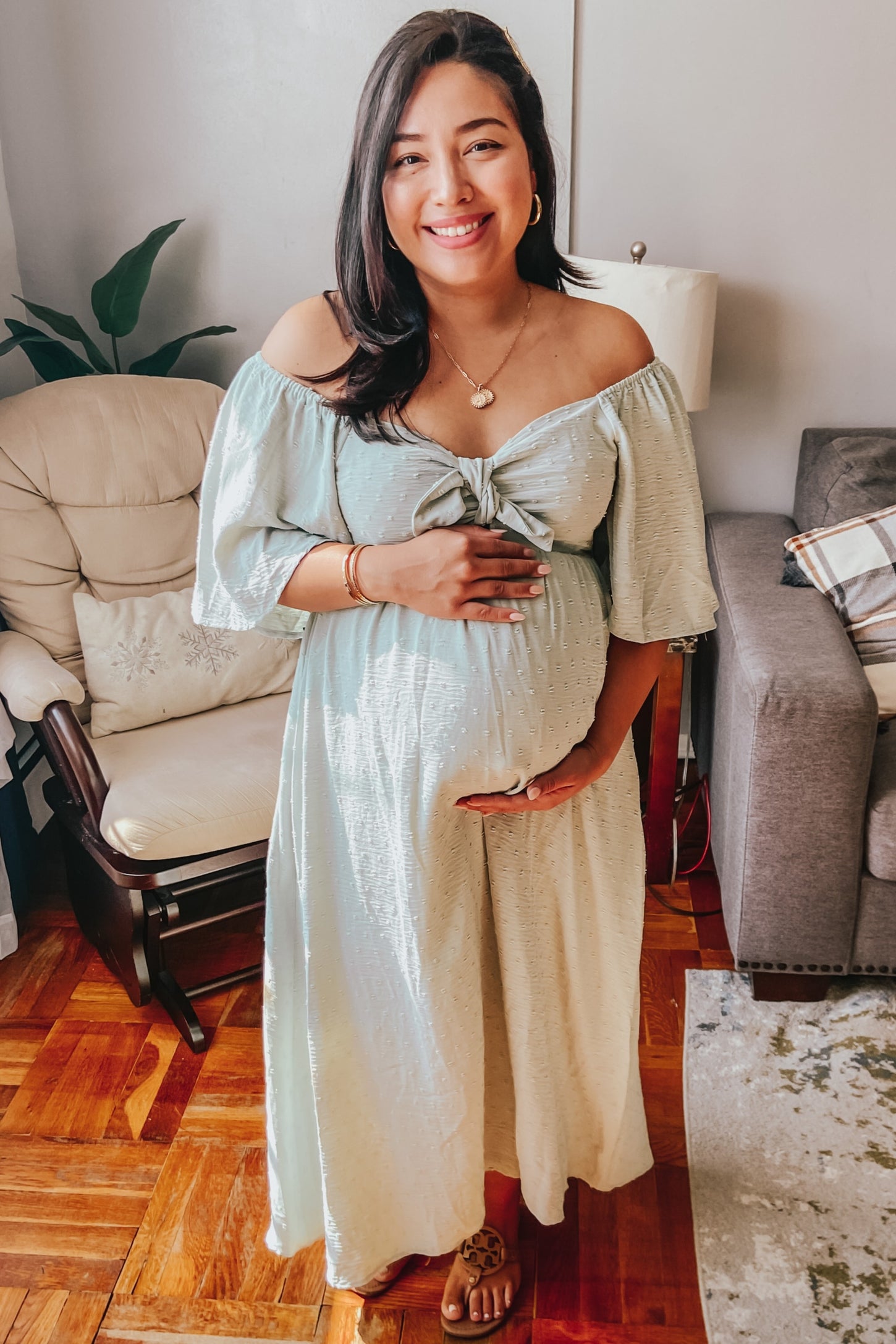 woman wearing a mint green off-the-shoulder maternity dress with a bow detail, holding her baby bump
